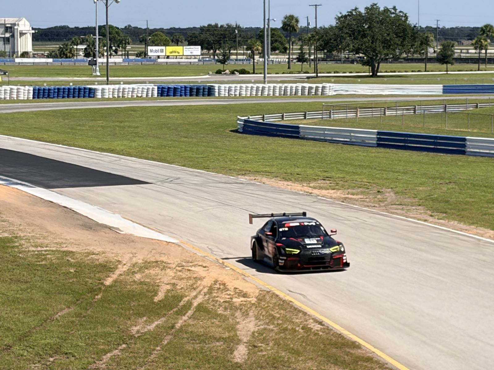 Black race car with a rear wing and green accents coming through a corner at Sebring