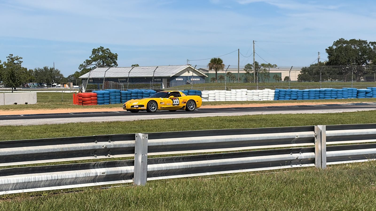 Yellow race car number 33 on track behind the guardrail, with blue and white tire barriers and a pit building in the background